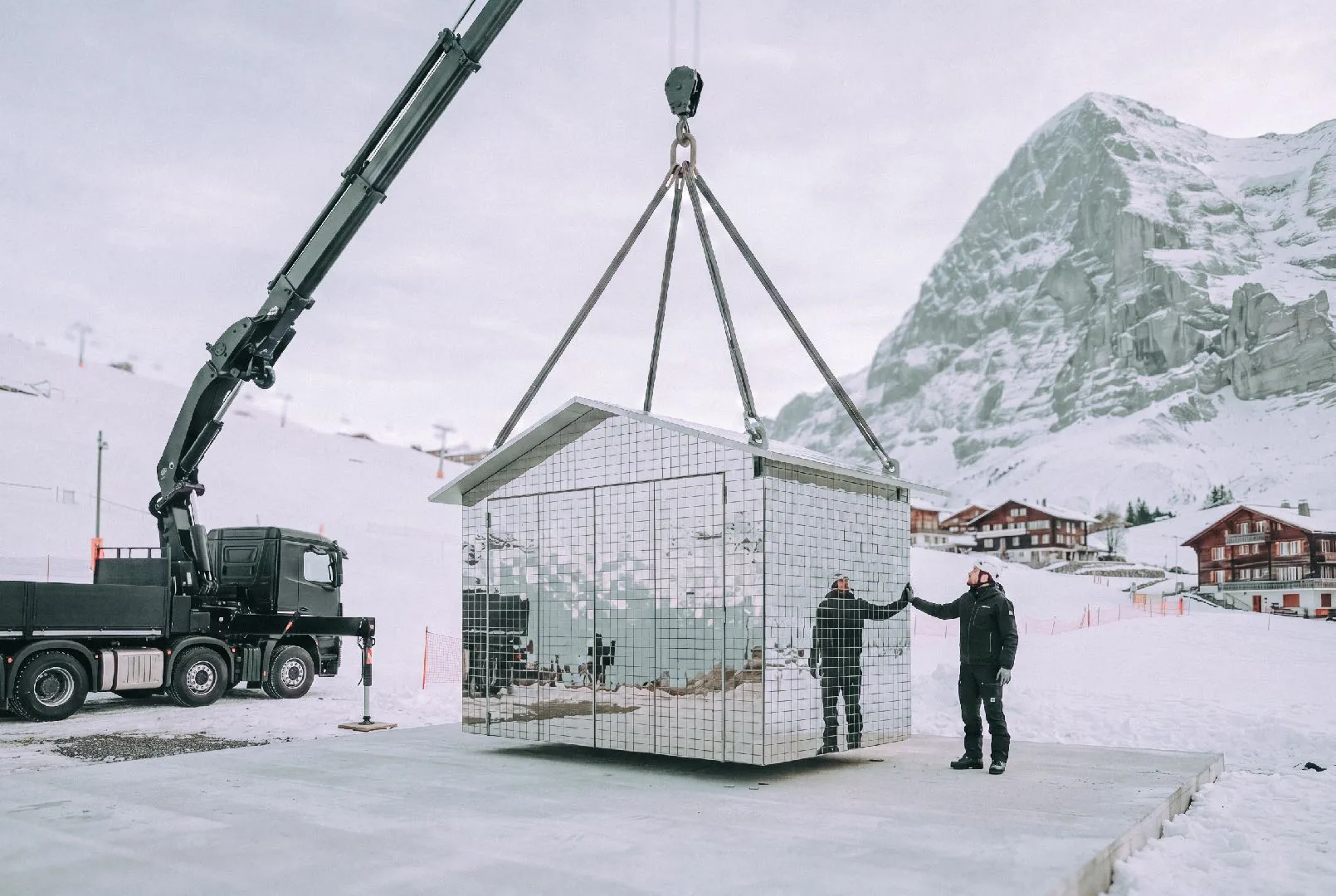 A mirrored pavilion is lifted by a truck crane onto a snowy plateau beneath alpine peaks